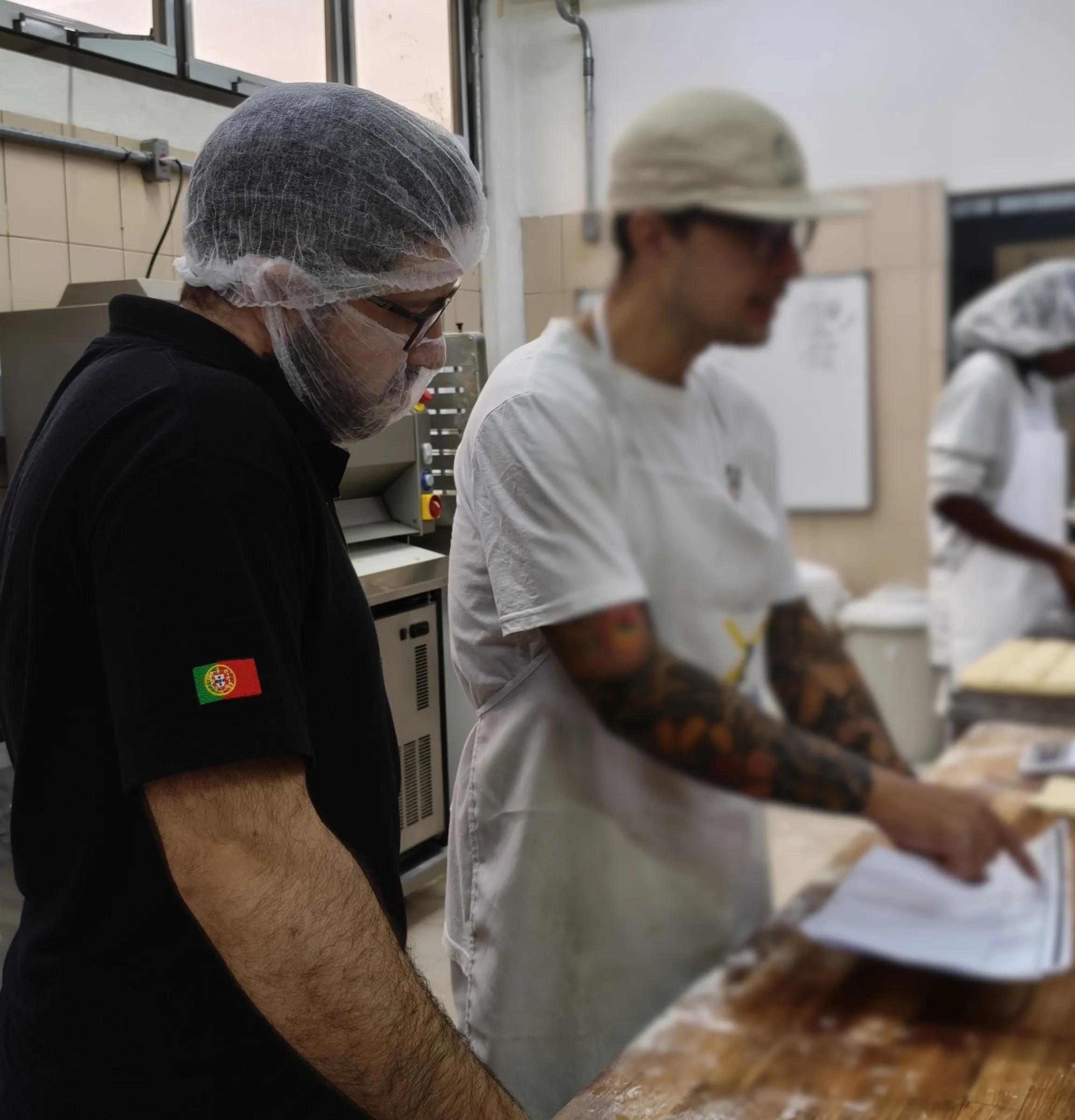 Padeiro conferindo um pedido impresso na bancada da produção ao lado de um colega de touca