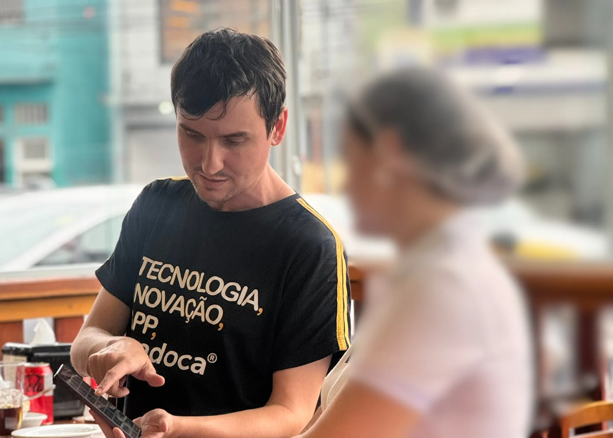 Homem com camiseta da Epadoca mostrando o celular para uma funcionária de touca em uma padaria