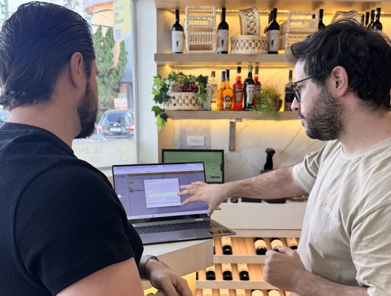 Dois homens no balcão de um estabelecimento, um deles apontando para a tela de um notebook