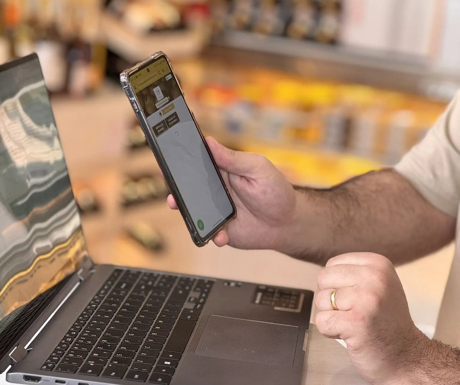 Mãos segurando um celular com a tela de pedidos aberta, ao lado de um notebook