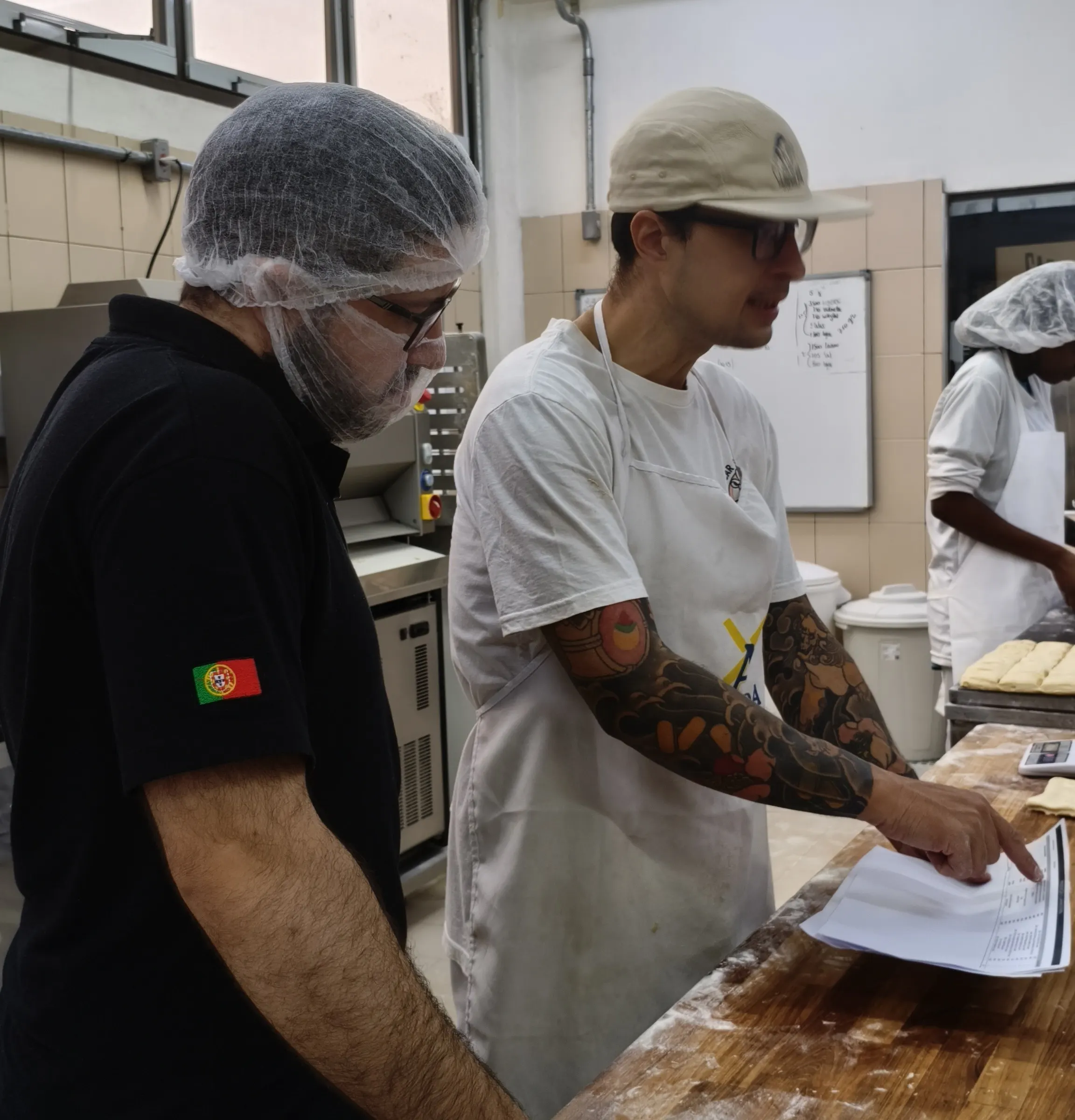 Padeiro conferindo um pedido impresso na bancada da produção ao lado de um colega de touca