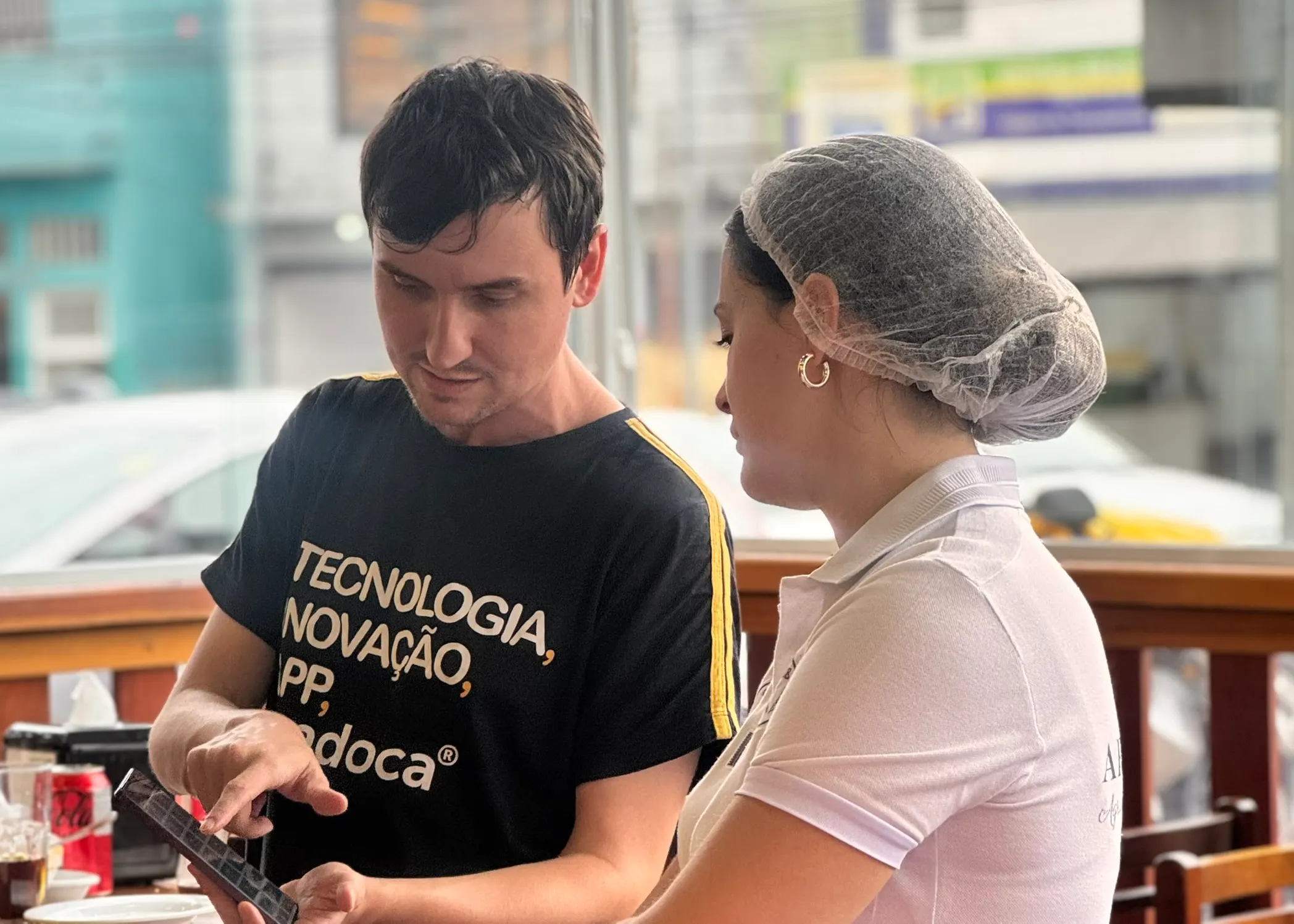 Homem com camiseta da Epadoca mostrando o celular para uma funcionária de touca em uma padaria